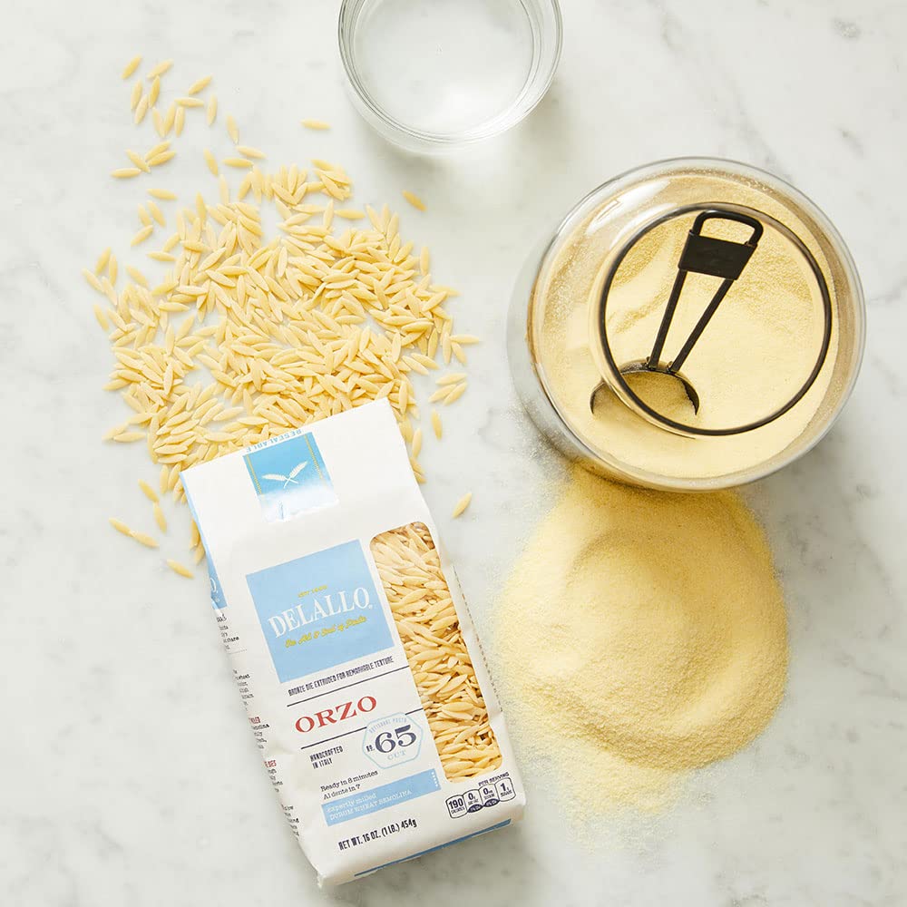 Orzo pasta packaging with uncooked orzo, a container of cooked orzo, and a spoon on a marble surface.
