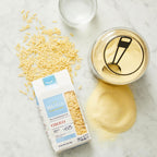 Orzo pasta packaging with uncooked orzo, a container of cooked orzo, and a spoon on a marble surface.