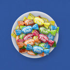 A white bowl filled with wrapped Swedish Fish candies in many bright colors, photographed from above against a solid blue background.
