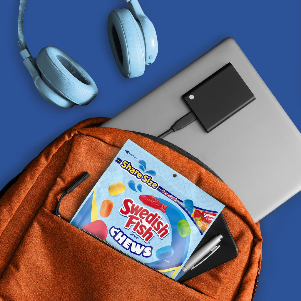 An orange backpack on a blue background, shown with a laptop, headphones, and accessories. A pack of Swedish Fish Chews is tucked into the front pocket.