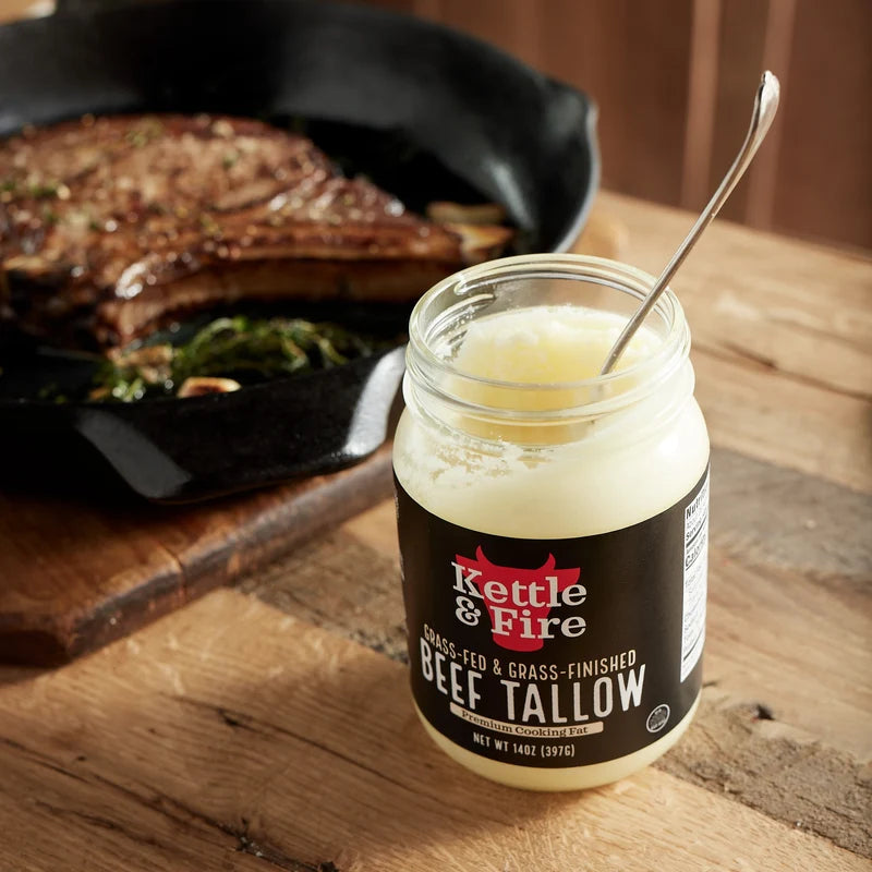 A jar of Kettle & Fire Beef Tallow lard oil with a metal spoon, set on a rustic wooden table with a cast iron skillet of cooked meat in the background.